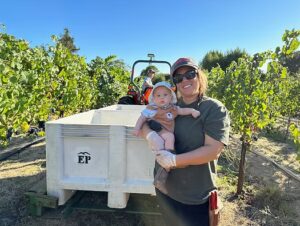 Woman in winery, holding baby. In background is farm machinery and vineyards.