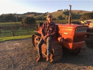 Man sitting on an orange crawler tractor in front of vineyard rows, with rolling green hills in the background.