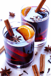 Two clear glasses of a warm, spiced beverage garnished with orange slices, cinnamon sticks, and star anise.