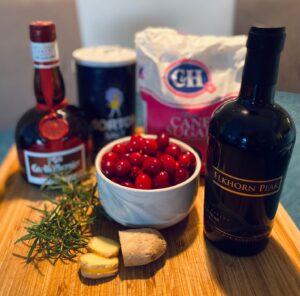Ingredients on a wooden board including cranberries, rosemary, ginger, salt, sugar, and Elkhorn Peak dessert wine.