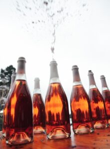 Several bottles of sparkling rosé on a wooden table, with one open and liquid spraying upward into the air.
