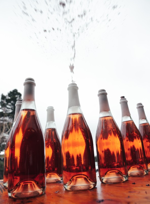 Several bottles of sparkling rosé on a wooden table, with one open and liquid spraying upward into the air.